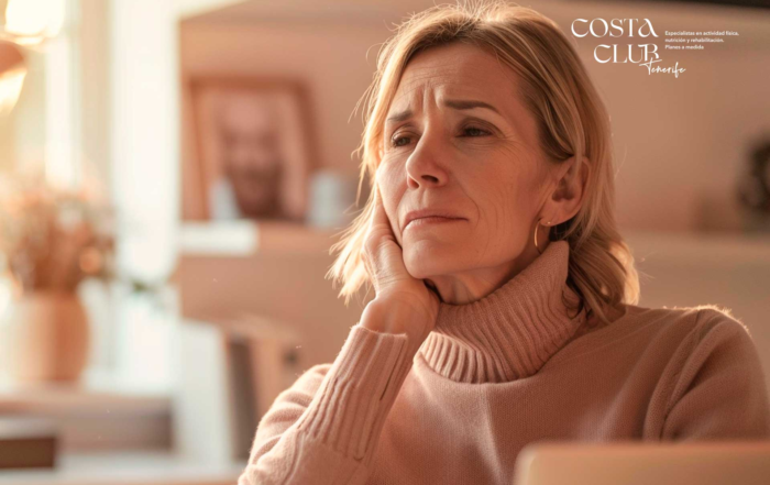 dolores-de-cuello - Costa Club Tenerife Older woman in a pink turtleneck sweater rests her chin on her hand with a thoughtful expression in an indoor setting; Costa Club Tenerife logo appears in the top right.