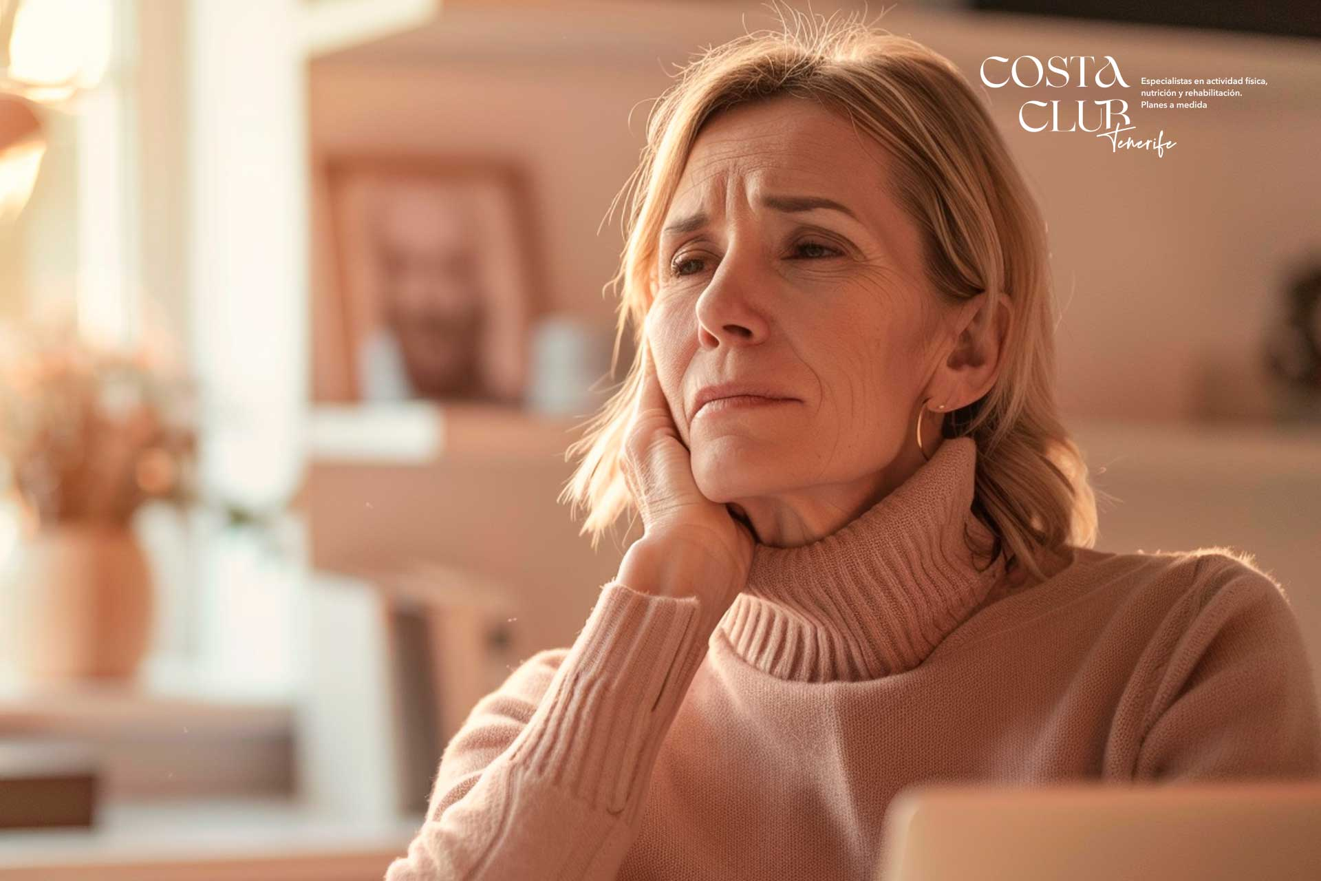dolores-de-cuello - Costa Club Tenerife Older woman in a pink turtleneck sweater rests her chin on her hand with a thoughtful expression in an indoor setting; Costa Club Tenerife logo appears in the top right.